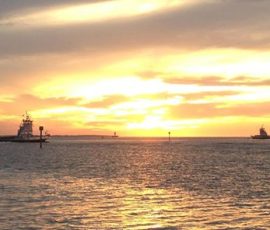 Two boats sail on calm water at sunset, with golden clouds lighting up the sky above the horizon.