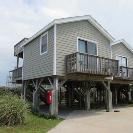 Elevated gray beach house with a balcony, wooden stilts, and parking space underneath, next to shrubs and concrete.