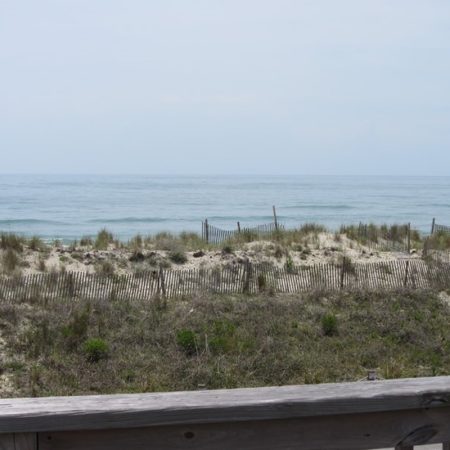 View of a sandy beach with grass, wooden fencing, and the ocean under a cloudy sky, seen from a wooden deck.