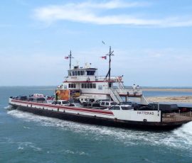A ferry named Hatteras transports vehicles and passengers across the water with a sandy shoreline in the background.