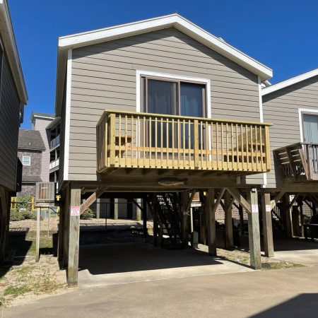 Gray stilt house with a wooden balcony and open parking space underneath, set against a clear blue sky.