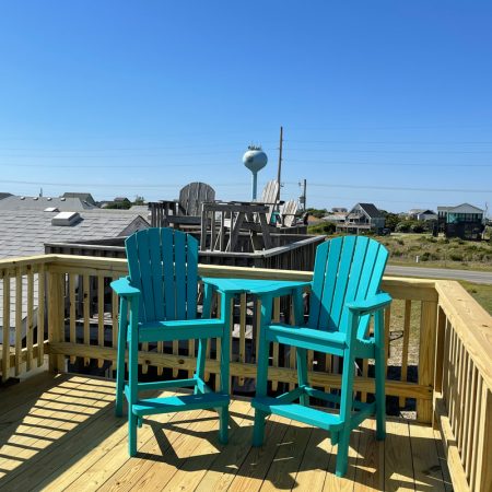 Two turquoise chairs and a table on a wooden deck overlook rooftops, a water tower, and a clear blue sky.