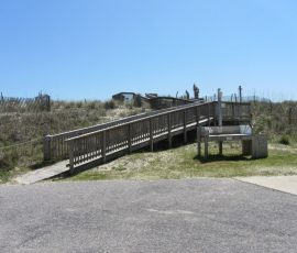 Wooden ramp leading up a sandy dune, fenced on both sides, under a clear blue sky.