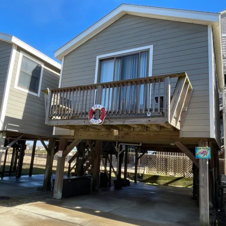 Beige stilt beach house with a wooden balcony, sliding glass doors, and a parking area underneath, on a sunny day.