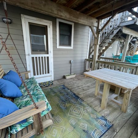 Wooden porch with a bench swing, table with benches, patterned rug, and stairs leading to an upper level.