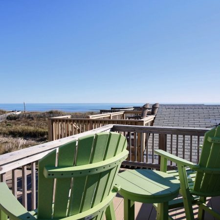 Two green chairs on a wooden deck overlook rooftops, dunes, and the ocean under a clear blue sky.