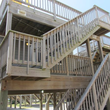 Wooden stairs and railings lead up to a multi-level elevated house on stilts, seen from an upward angle.