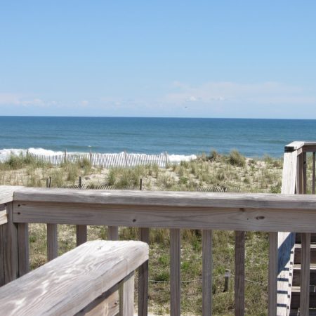 Wooden deck overlooking grassy dunes, a white sand fence, and calm ocean waves under a clear blue sky.