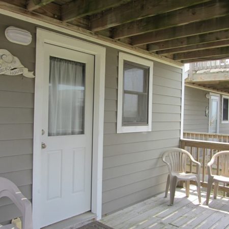 Gray house porch with three beige plastic chairs, a white door, two windows, and a “Welcome” sign on the wall.