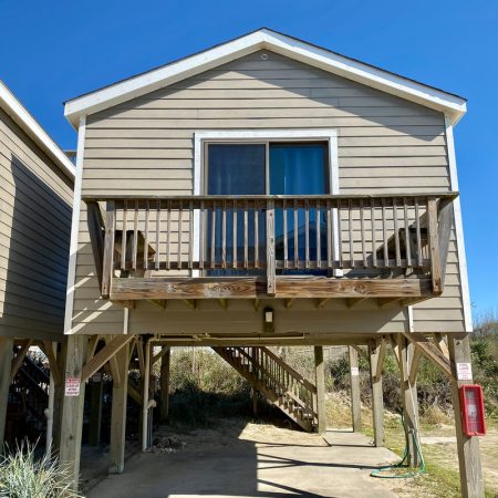 Elevated beach house with a small wooden balcony, staircase, and sandy plants in the background under a clear blue sky.