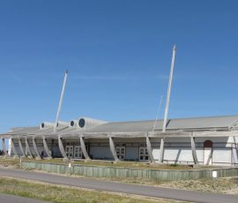 Long, low white building with a curved metal roof and tall poles in front, set beside a road under a clear blue sky.