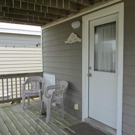 Two plastic chairs sit on a wooden porch next to a white door with a curtain, under a covered roof.