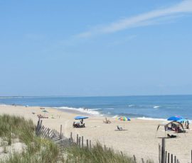 Sandy beach with scattered people, umbrellas, ocean waves, and grassy dunes under a clear blue sky.