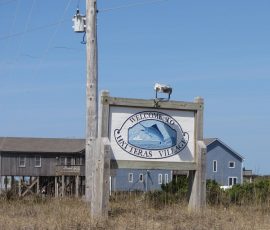 A wooden sign reads “Welcome to Hatteras Village” with houses and dry grass in the background under a clear sky.