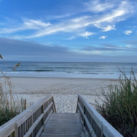 Wooden boardwalk leads to a sandy beach with ocean waves under a blue sky with scattered clouds.
