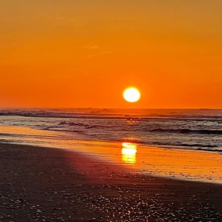 Sunset over the ocean with gentle waves, orange sky, and sunlight reflecting on the wet sandy beach.