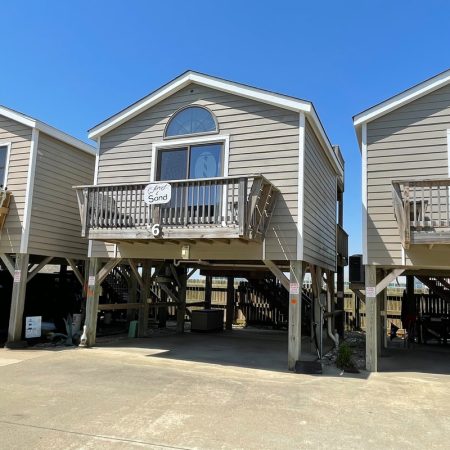 Raised beach house with a small balcony, labeled “Sea Sand,” flanked by similar homes under a clear blue sky.