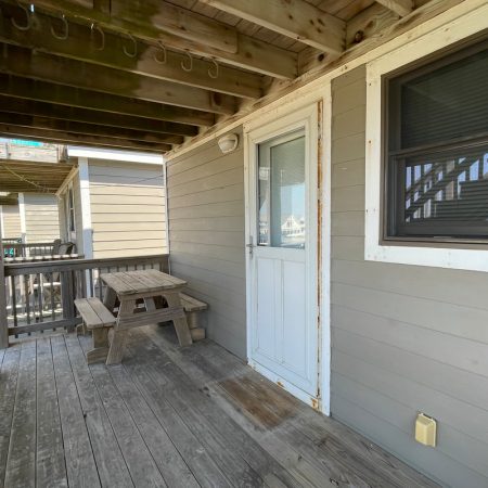 Wooden porch with a small picnic table, bench, door, window, and overhead beams, all in a neutral color scheme.