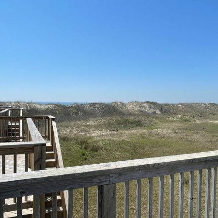 Wooden deck overlooking grassy sand dunes under a clear blue sky, with the ocean visible in the distance.