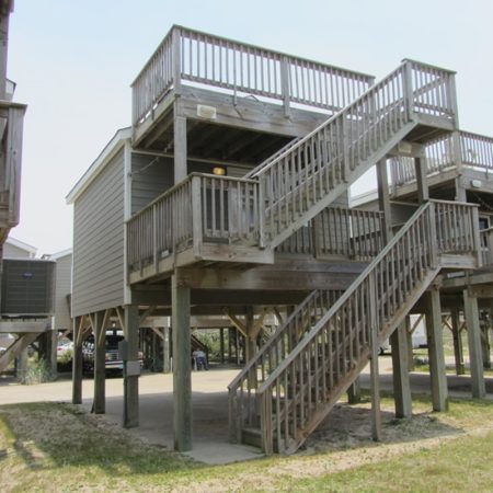 Elevated beach house on wooden stilts with two staircases leading to a deck and covered parking area below.