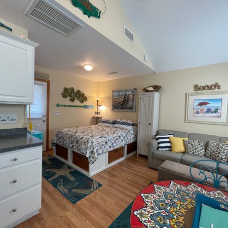 Small beach-themed studio room with a bed, sofa, kitchen area, and coastal decor accents on walls and furniture.