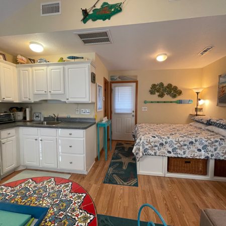 Small studio with white kitchen, bed with patterned bedding, wood floors, and coastal-themed decor.