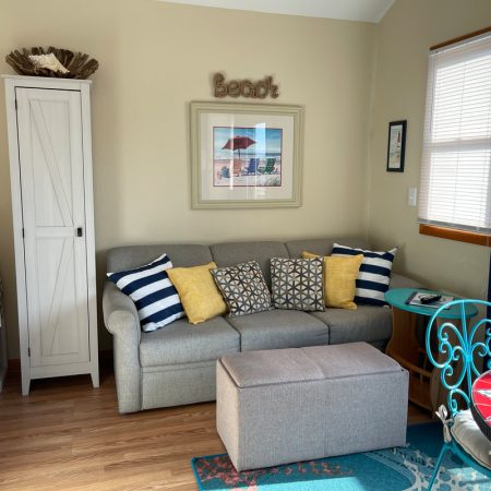 A small living room with a gray sofa, colorful pillows, an ottoman, and beach-themed decor.