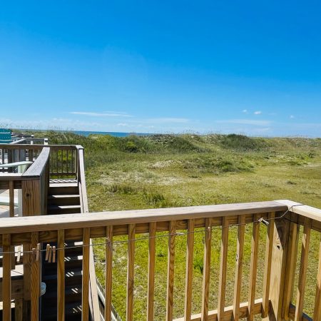 Wooden deck with chairs overlooks grassy sand dunes under a clear blue sky on a sunny day.