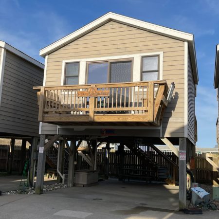 Elevated beige beach house with a wooden balcony and stairs, situated on concrete stilts, with clear sunny weather.