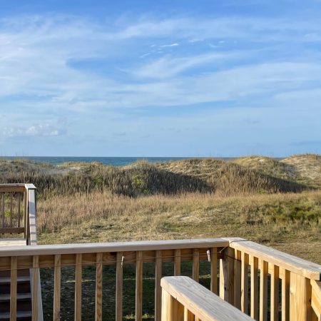 Wooden deck overlooks sandy dunes and grassy area, with a blue sky and ocean visible in the background.