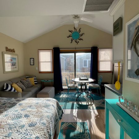 Sunny bedroom with a bed, sofa, dining table, pelican art, and beach-themed decor, overlooking a small balcony.