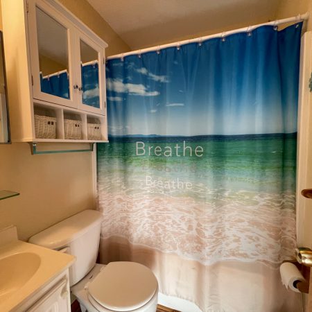 Small bathroom with a beach-themed shower curtain that reads “Breathe,” a toilet, sink, mirror cabinet, and wall shelf.