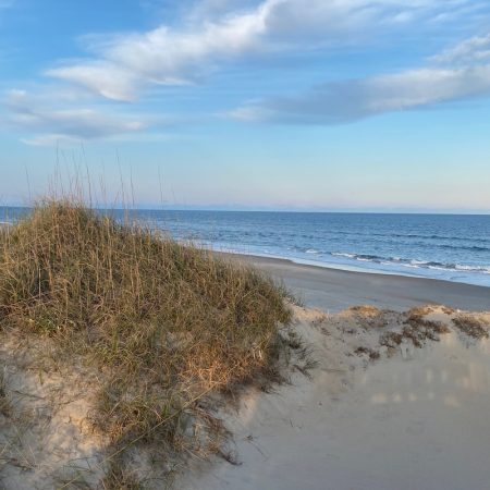 Sandy dunes with grass in the foreground, ocean waves, and a blue sky with scattered clouds in the background.