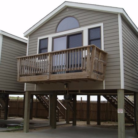 Beige stilt house with a small wooden balcony, large windows, and stairs leading up from the ground level.