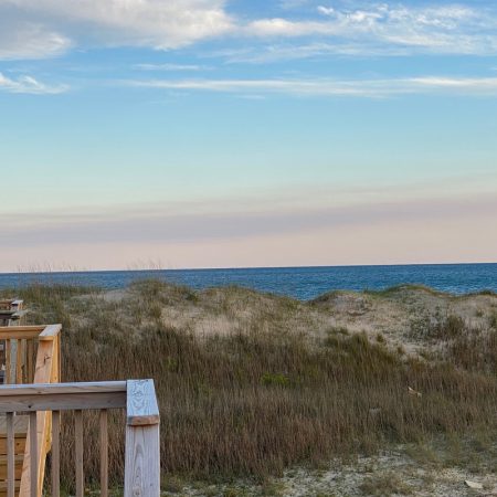 Wooden deck overlooking sandy dunes, grassy vegetation, and a calm ocean under a blue sky with scattered clouds.