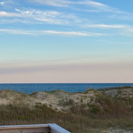 View of sand dunes and grassy vegetation with the ocean and sky in the background, seen from a wooden railing.