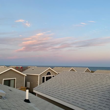 Rows of gray rooftops lead to a calm ocean horizon under a sky with soft pink clouds at sunset.