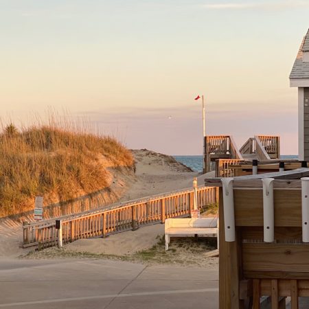 Wooden walkways lead over sand dunes to the beach, with a red flag and calm sea visible under a pastel sky.