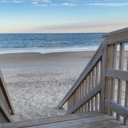 Wooden stairs lead down to a sandy beach with calm ocean waves under a partly cloudy sky.