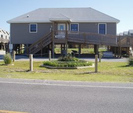 Elevated gray house with ramp and stairs, VACANCY sign, driveway, garden bed, and other houses in the background.
