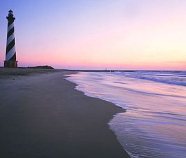 Black-and-white striped lighthouse stands on a sandy beach at sunset with calm waves and a pastel sky.