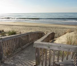 Wooden stairs lead down to a sandy beach with ocean waves and sparse grass under a clear sky.