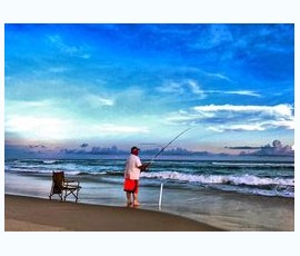 A person in red shorts fishes on a sandy beach near a chair, with waves and a blue sky in the background.