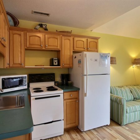 Small kitchenette with wooden cabinets, white appliances, microwave, sink, and a striped couch in a cozy room.