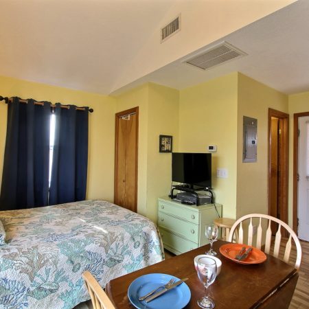 Small bedroom with a bed, TV on a dresser, dining table set for two, and a door with a window, all in natural light.