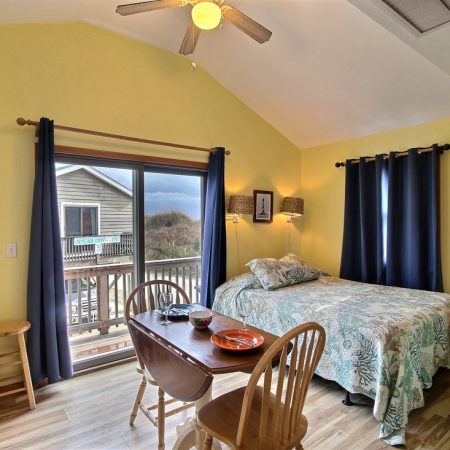 Cozy room with a bed, dining table, and balcony view; yellow walls, hardwood floor, and blue curtains.