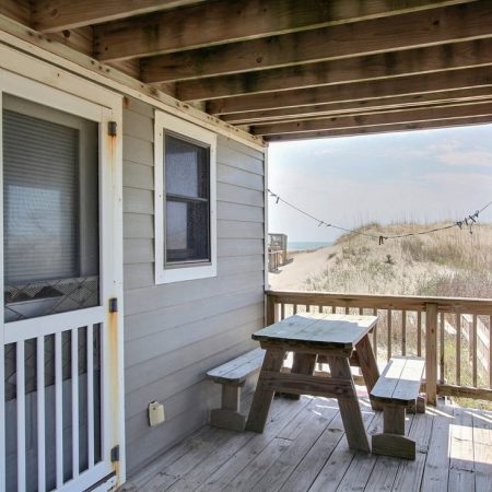 Wooden porch with a picnic table, overlooking sand dunes and a glimpse of the beach in the background.