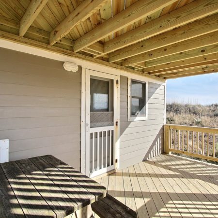 Covered wooden porch with picnic table, railing, and a door leading into a gray house. Sandy landscape in the background.