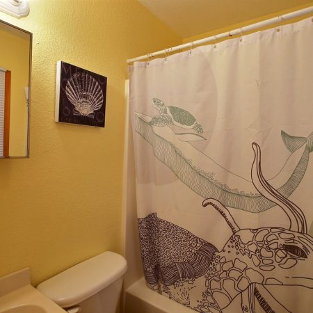 Small bathroom with yellow walls, a mirror, seashell art, and a shower curtain featuring sea turtles and ocean designs.
