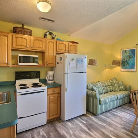 Small kitchen with wooden cabinets and appliances, next to a striped sofa, dining table, and large window with blue curtains.